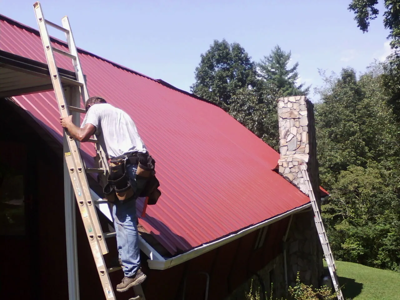 Roofer on a ladder beside a red metal roof and stone chimney — Soffit & Fascia Repair by Roofing Brothers in Hartsdale